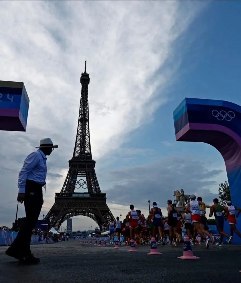 Race walkers pass the Eiffel Tower on the Paris 2024 Olympic course as an official in a white hat watches from the roadside.